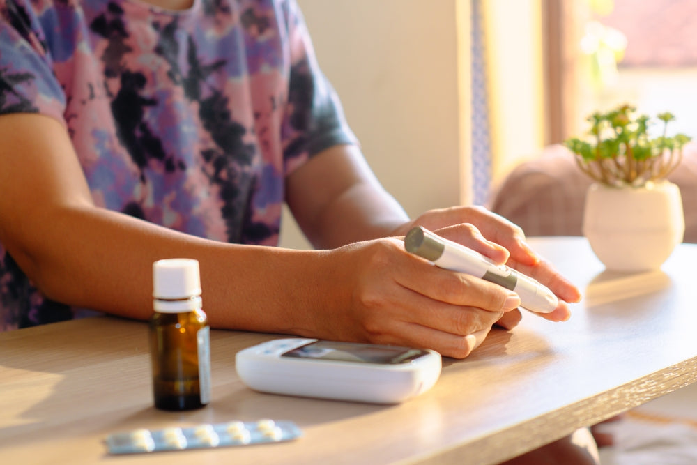 Person using lancet pen for blood sugar test with glucometer, medicine bottle, and pills on wooden table, diabetes healthcare concept.